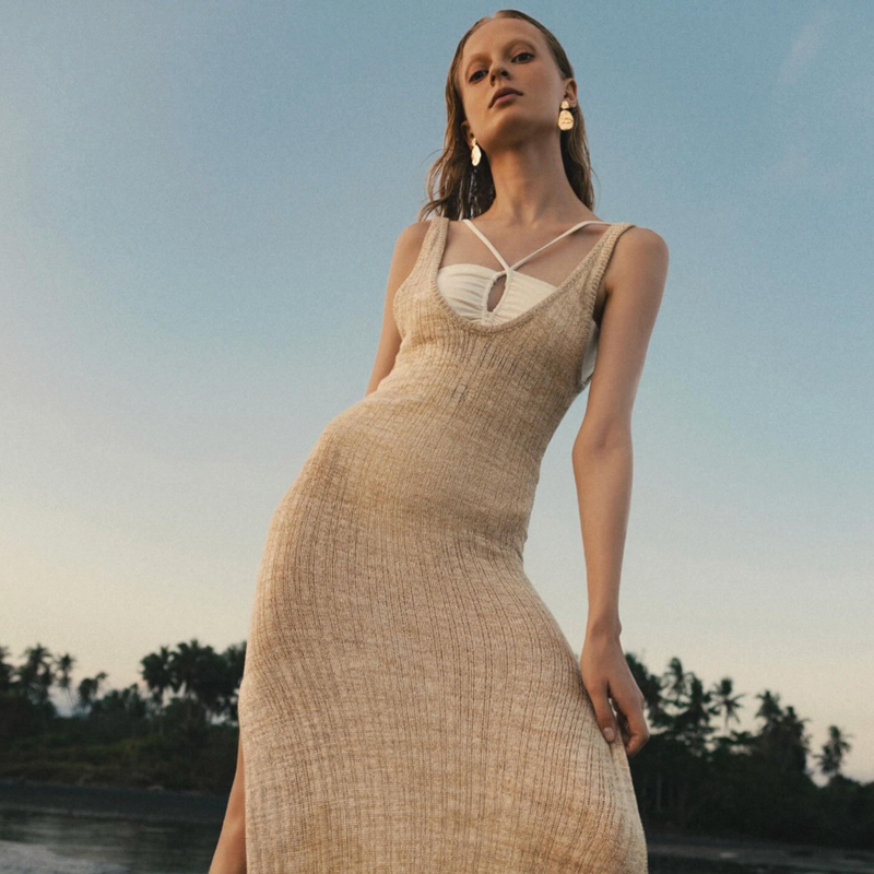 Woman in a beige dress standing against a scenic background with trees and water.